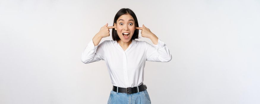 Young annoyed asian girl close ears and screaming, fed up with loud noise, terrible music, standing over white background. Copy space