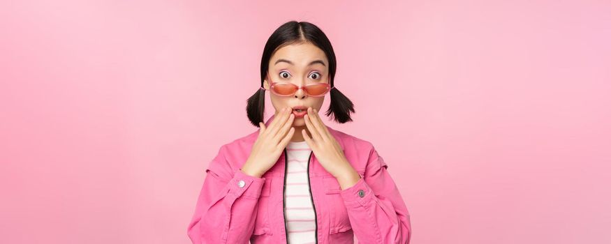 Portrait of young asian woman in stylish sunlgasses, looking surprised, disbelief reaction, standing over pink background. Copy space