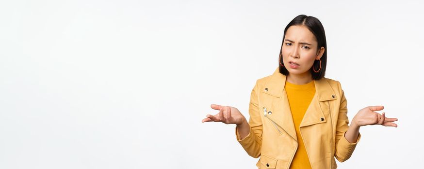 Image of confused young asian woman shrugging shoulders, looking puzzled and clueless at camera, cant understand, standing against white background.
