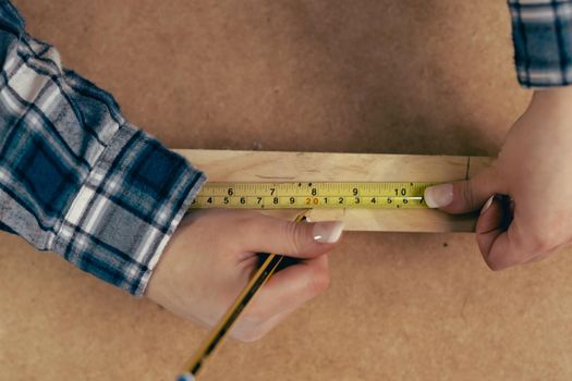 Top view of the hands of a young woman measuring a strip of wood for the manufacture of a piece of furniture in her small workshop, dressed in a blue checked shirt and red t-shirt. Young businesswoman handcrafting a piece of wood in her small business. Warm light indoors, background with wooden slats.