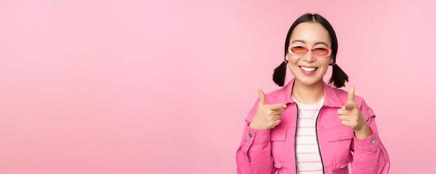 Close up portrait of modern asian girl in sunglasses smiling, pointing fingers at camera, praise you, inviting or complimenting, standing over pink background.