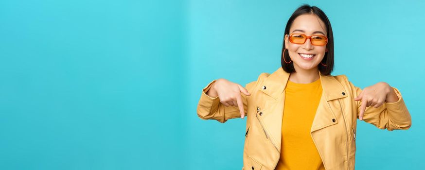 Portrait of stylish asian girl, wears sunglasses, smiles and points fingers down, shows advertisement, stands over blue background.