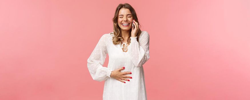 Portrait of joyful happy bright young girl in white dress, having fun, speaking to friend, calling person mobile phone, laughing and touching belly, close eyes smiling, hold smartphone near ear.
