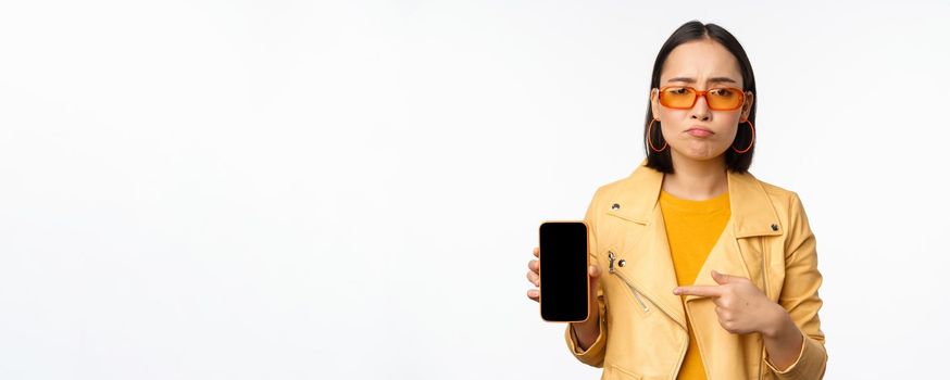 Portrait of sad asian woman in sunglasses, pointing finger at mobile phone app interface, showing smartphone application, standing over white background.