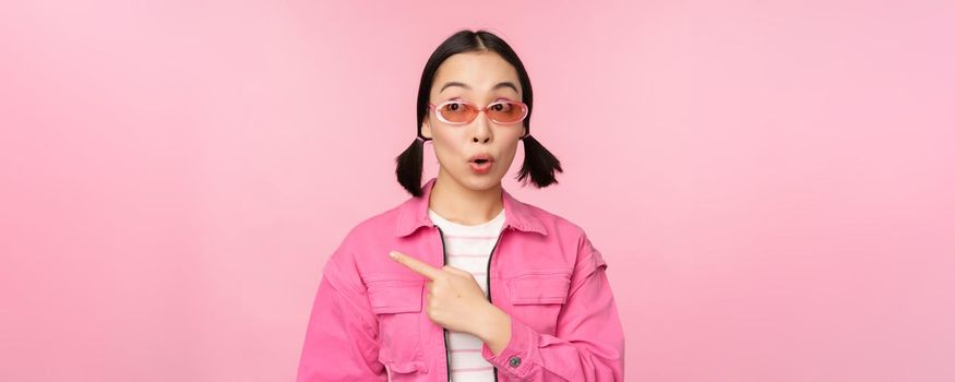 Portrait of smiling asian girl in stylish outfit, sunglasses, pointing finger left, showing advertisement, banner, standing over pink background. Copy space