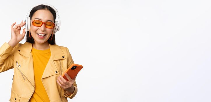 Happy stylish asian woman in sunglasses, listening music in headphones, holding smartphone, singing and dancing, standing over white background.