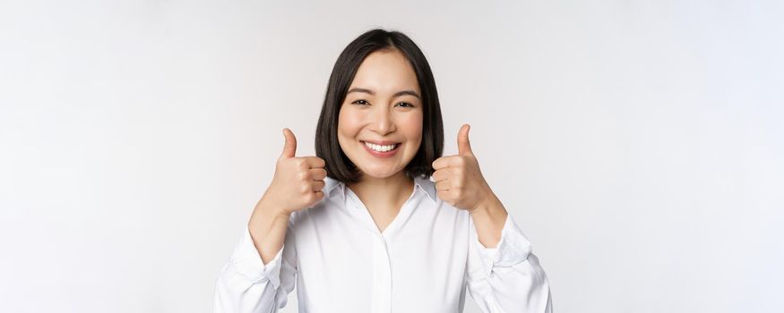 Cheerful asian woman face, showing thumbs up and smiling, pleased by something, recommending smth good, standing over white background.