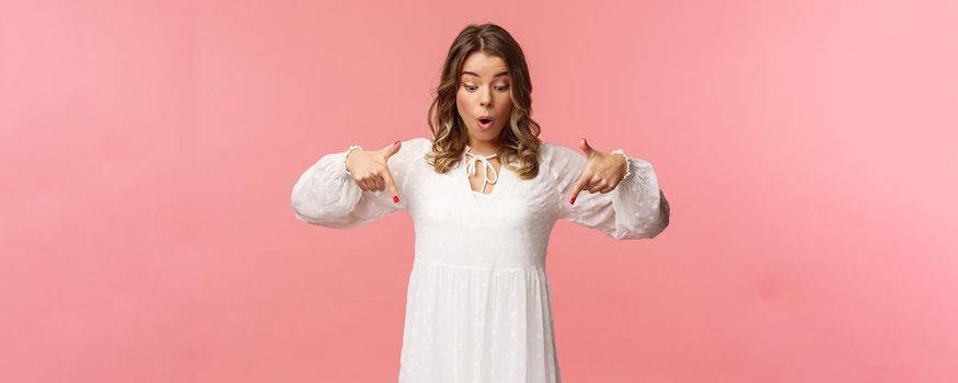 Amazed and wondered good-looking blond european woman 20s, wear beautiful white dress, looking and pointing up, freaking out over something awesome and breathtaking, pink background.