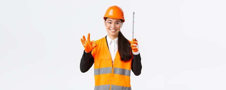Satisfied asian female construction engineer, architect or inspector at enterprise showing okay gesture and tape measure, smiling pleased, give permission, approve measurements are valid.