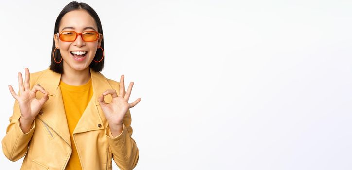 Enthusiastic asian girl in sunglasses, shows okay, ok sign in approval, smiling and laughing, approve, recommend smth, praise and compliment, standing over white background.