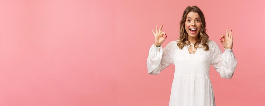 Portrait of happy and pleased good-looking blond woman in white dress, recommend great product, leave positive review, show okay signs and smiling satisfied, standing pink background.