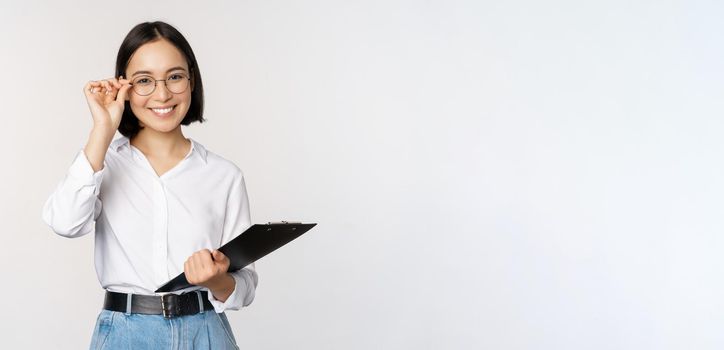 Young woman, office worker manager in glasses, holding clipboard and looking like professional, standing against white background.