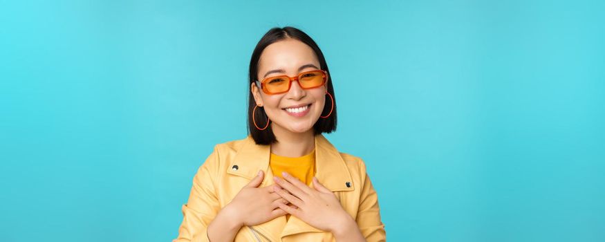 Close up portrait of asian young woman in sunglasses, smiling and looking romantic, standing happy over blue background. Copy space