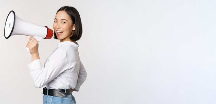 Beautiful young asian woman talking in megaphone, screams in speakerphone and smiling, making announcement, shout out information, standing over white background.