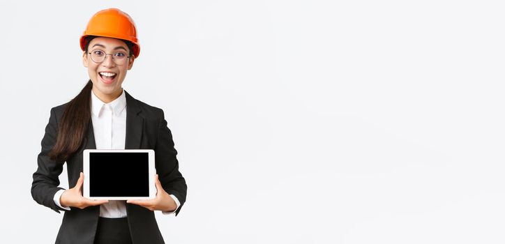 Excited and amazed smiling asian female construction engineer, architect in safety helmet and business suit, showing digital tablet screen with amazed expression, standing white background.