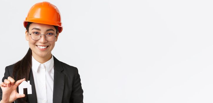 Close-up of happy asian female engingeer, real estate agent in helmet and business suit holding house miniature and smiling, architect working over renovation project, white background.