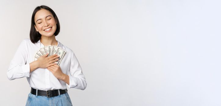 Happy asian woman hugging money dollars and smiling satisfied, standing over white background. Copy space