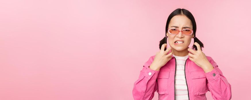 Image of angry, pissed off korean adult female model, shaking fists and shouting, screaming outraged, standing over pink background.