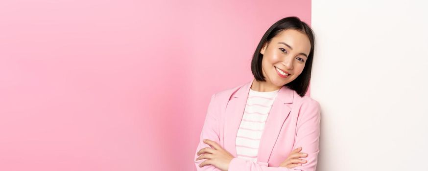 Confident, successful japanese office lady in suit, cross arms, looking as professional at camera, leaning on white wall with advertisement, empty copy space for logo, pink background.