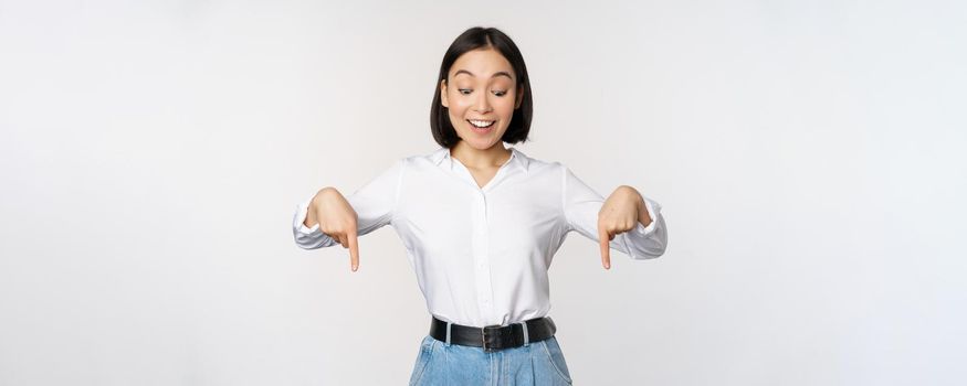 Portrait of happy asian woman pointing fingers down and looking below at advertisement, showing info banner, advertising, standing over white background.