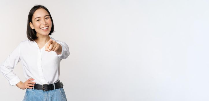 Its you. Beautiful young asian woman, company manager pointing finger at camera and smiling, choosing, inviting people, recruiting, standing over white background.