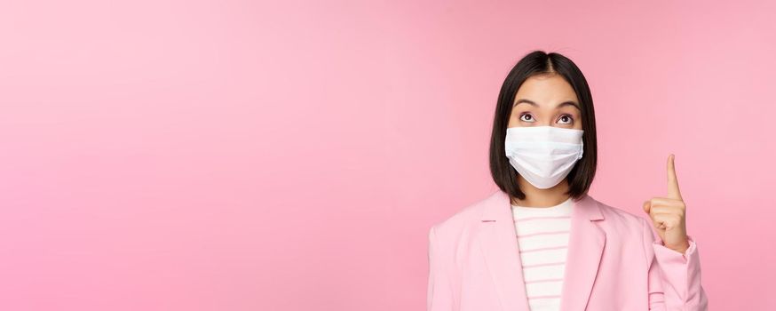 Close up portrait of businesswoman, asian lady in office suit and medical face mask, looking and pointing up, showing company logo or banner on top, pink background.