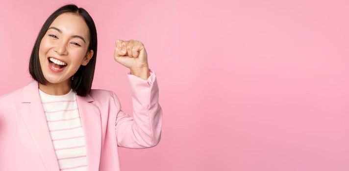 Enthusiastic asian corporate woman, businesswoman raising hand up and cheering, triumphing, winning and celebrating, standing over pink background.