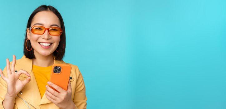 Happy korean girl in sunglasses, showing okay sign and holding mobile phone, using smartphone app, recommending application, standing over blue background.
