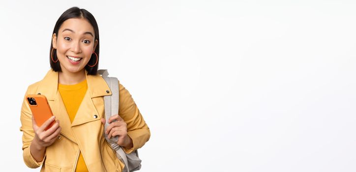 Image of happy girl traveller, tourist with backpack looking at smartphone, using mobile phone route app, standing over white background. Copy space