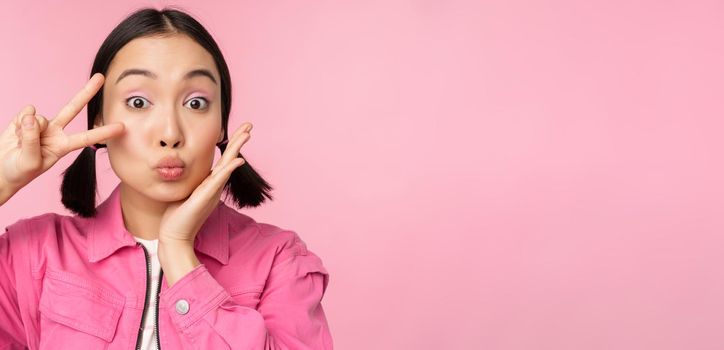 Close up portrait of stylish asian girl shows peace, v-sign and kissing, posing against pink background.