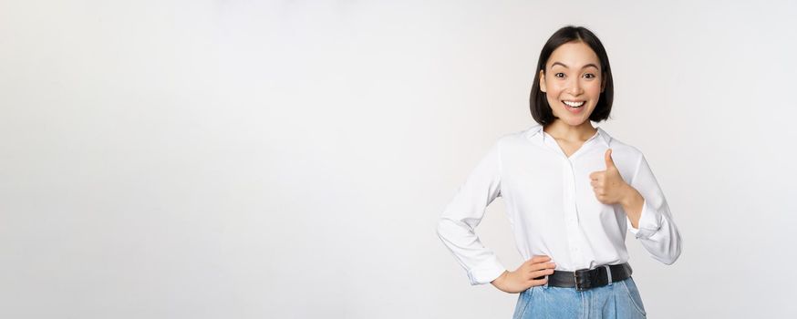 Very good. Smiling enthusiastic asian female entrepreneur, showing thumb up in approval, praise excellent job, satisfied with results, standing over white background.