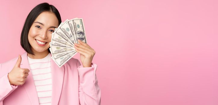 Microcredit, investment and business people concept. Young asian businesswoman, corporate lady showing money, cash dollars, thumbs up, recommending company, pink background.