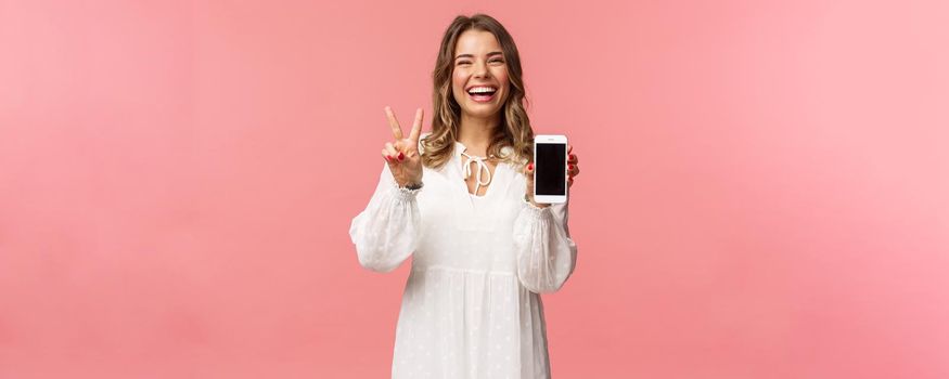 Portrait of kawaii optimistic and happy young girl in white dress, show mobile phone display and peace sign, laughing feeling cheerful and glad to share awesome app, useful link, pink background.
