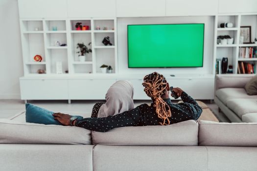 African Couple Sitting On Sofa Watching TV Together Chroma Green Screen Woman Wearing Islamic Hijab Clothes. High quality photo