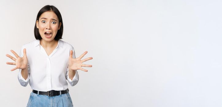 Asian woman looks at camera and screams in panic. Young korean girl looking anxious, panicking, shaking hands and shouting, standing over white background.