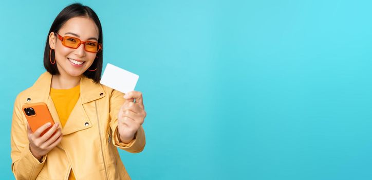 Online shopping. Stylish young asian woman in sunglasses, showing credit card and using smartphone, paying in internet, making purchase, standing over blue background.