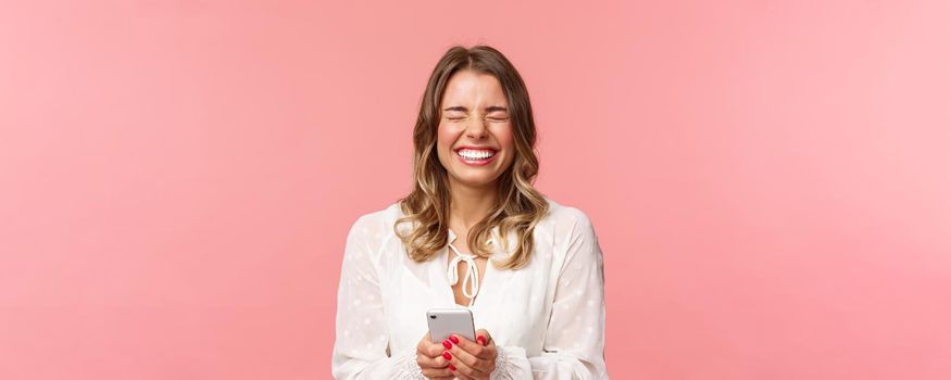 Close-up portrait of carefree tender, lovely blond girl in white dress, laughing over funny joke or message, holding mobile phone, close eyes and giggle at something hilarious, pink background.