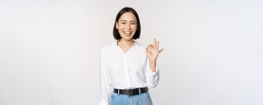 Very good. Smiling asian woman showing okay with satisfied face expression, praise and compliment great job, pleased by smth, standing over white background.