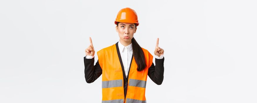 Building, construction and industrial concept. Disappointed gloomy asian female engineer, architect in safety helmet and reflective clothing pouting displeased as looking and pointing fingers up.