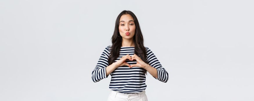 Lifestyle, people emotions and casual concept. Lovely smiling adorable asian woman showing heart sign and smiling, express sympathy or care, sending air kiss at camera, standing white background.