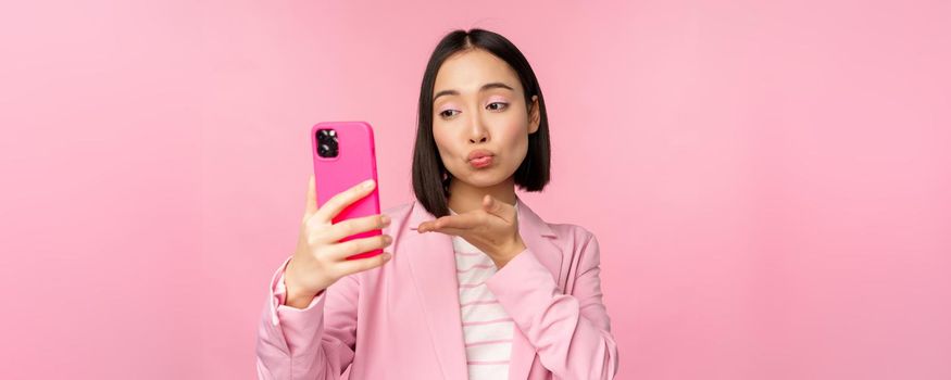 Stylish asian businesswoman, girl in suit taking selfie on smartphone, video chat with mobile phone app, posing against pink studio background.