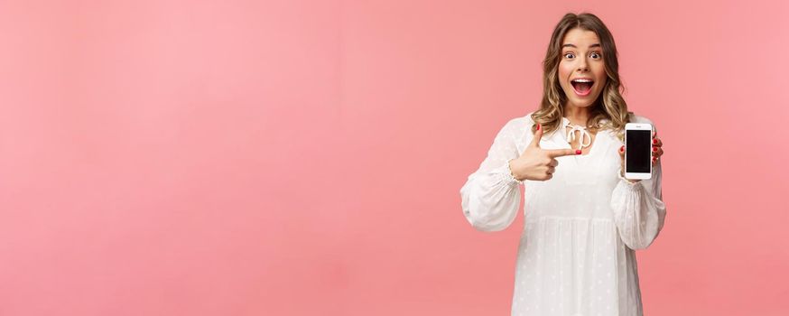 Portrait of impressed, excited young blond woman showing something awesome on display, pointing mobile phone screen and smiling astonished, brag with her recent match on dating app.