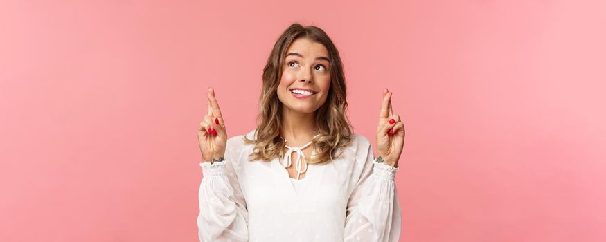 Close-up portrait of hopeful, optimistic attractive woman in white dress, bite lip and smiling as daydreaming, hope dream come true, making wish or praying, standing pink background.