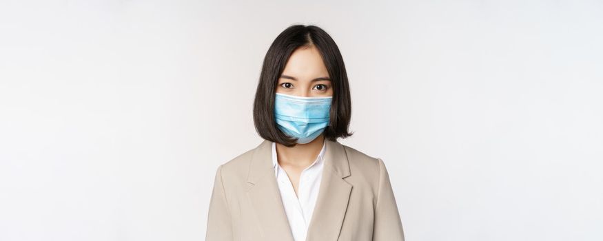 Coronavirus and workplace concept. Portrait of asian office worker, businesswoman in medical face mask looking at camera, white background.