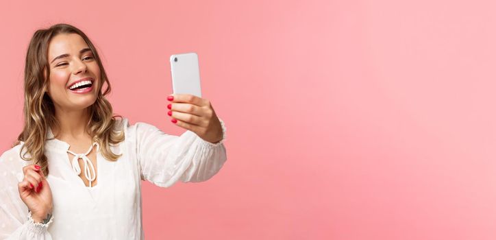 Close-up portrait of cheerful upbeat smiling blond girl, wearing white dress, laughing as record video, calling friend on mobile application, taking photo, selfie on smartphone, pink background.