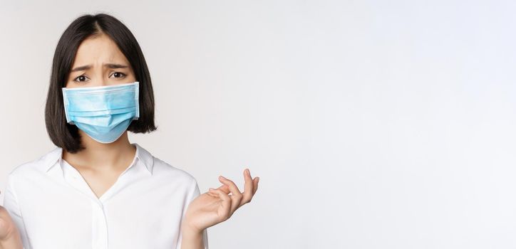 Distressed and miserable young asian woman in face mask, looking up, looking up sad, standing over white background.