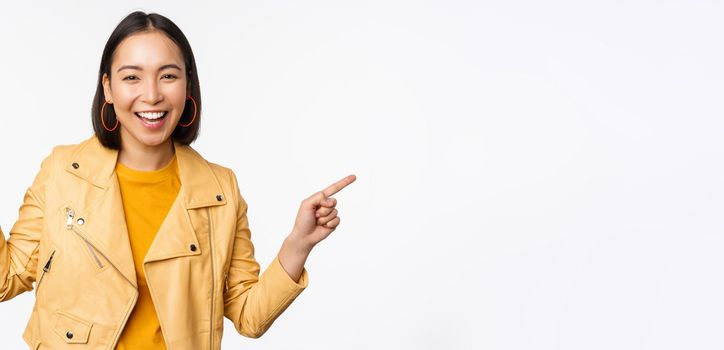 Happy beautiful asian girl pointing fingers left and right, showing banner logo, demonstrating sale, standing in yellow jacket over white background.