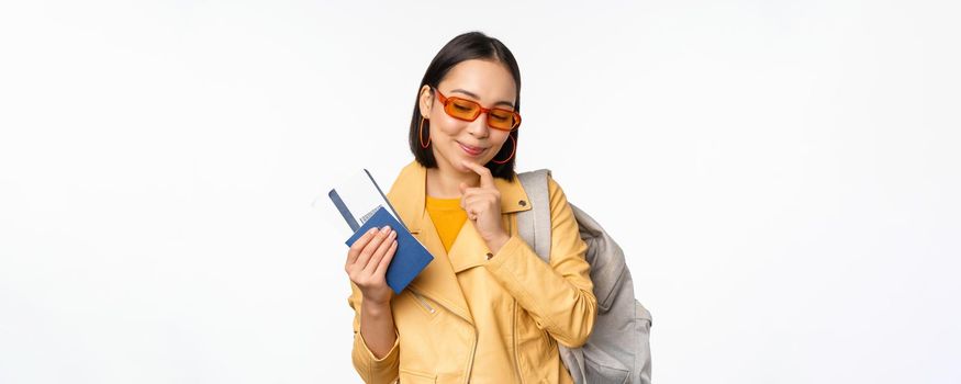 Asian girl tourist with boarding tickets and passport going abroad, holding backpack, thinking of travelling, standing over white background.