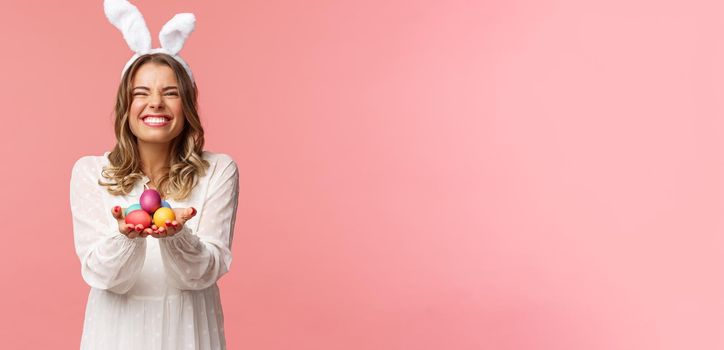 Holidays, spring and party concept. Portrait of excited charming blond woman with lovely pleased smile, holding painted eggs, wear Easter day outfit, rabbit ears and white dress, pink background.