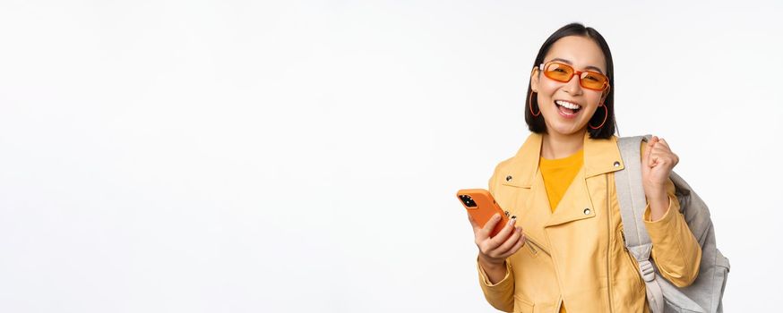 Young asian woman traveller, girl tourist in sunglasses, holding backpack and mobile phone, using application on smartphone, standing over white background.